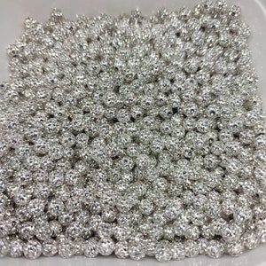 May include: A close-up view of a container filled with silver-colored beads. The beads are round and have a textured surface.