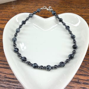 May include: A black beaded bracelet with snowflake obsidian and hematite beads. The bracelet is on a white heart-shaped dish, with a silver clasp. The beads are approximately 0.25 inches in diameter.