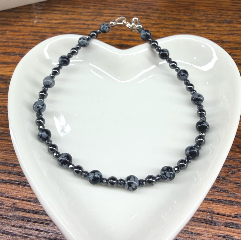 May include: A black beaded bracelet with snowflake obsidian and hematite beads. The bracelet is on a white heart-shaped dish, with a silver clasp. The beads are approximately 0.25 inches in diameter.