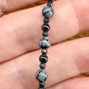 May include: A beaded bracelet featuring a mix of black and gray snowflake obsidian and hematite beads. The bracelet's design includes round and faceted beads, strung together with a silver-colored clasp.