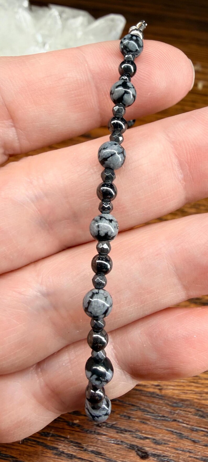 May include: A beaded bracelet featuring a mix of black and gray snowflake obsidian and hematite beads. The bracelet's design includes round and faceted beads, strung together with a silver-colored clasp.