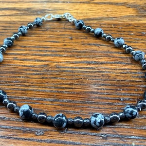 May include: A beaded bracelet featuring black and white speckled stones and smaller black beads. The bracelet is strung on a silver-colored chain and clasp. The jewelry is displayed on a wooden surface.