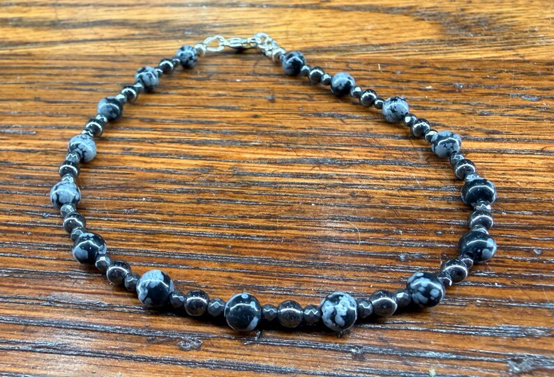 May include: A beaded bracelet featuring black and white speckled stones and smaller black beads. The bracelet is strung on a silver-colored chain and clasp. The jewelry is displayed on a wooden surface.