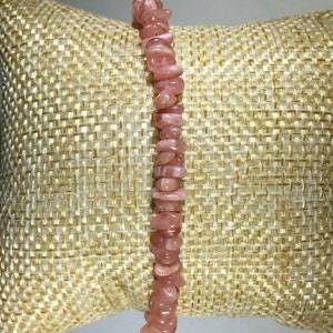 May include: A pink gemstone bracelet composed of small, irregular-shaped stones. The bracelet is displayed on a textured, beige surface, showcasing the natural color variations of the stones. The bracelet is a delicate piece of jewelry.