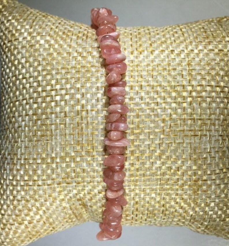 May include: A pink gemstone bracelet composed of small, irregular-shaped stones. The bracelet is displayed on a textured, beige surface, showcasing the natural color variations of the stones. The bracelet is a delicate piece of jewelry.