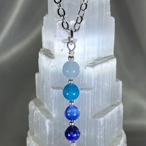 May include: A silver chain necklace with a pendant featuring a gradient of blue and white gemstone beads. The beads are arranged in a vertical line, transitioning from light blue to deep indigo. The necklace is displayed against a textured white background.