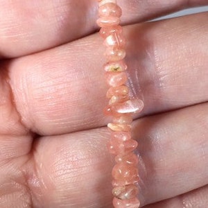 May include: A delicate bracelet composed of small, irregular, peach-colored gemstone beads. The beads are strung together, creating a flexible strand. The bracelet is held in a hand, showcasing its color and texture.