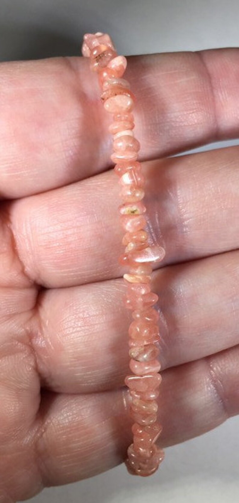 May include: A delicate bracelet composed of small, irregular, peach-colored gemstone beads. The beads are strung together, creating a flexible strand. The bracelet is held in a hand, showcasing its color and texture.