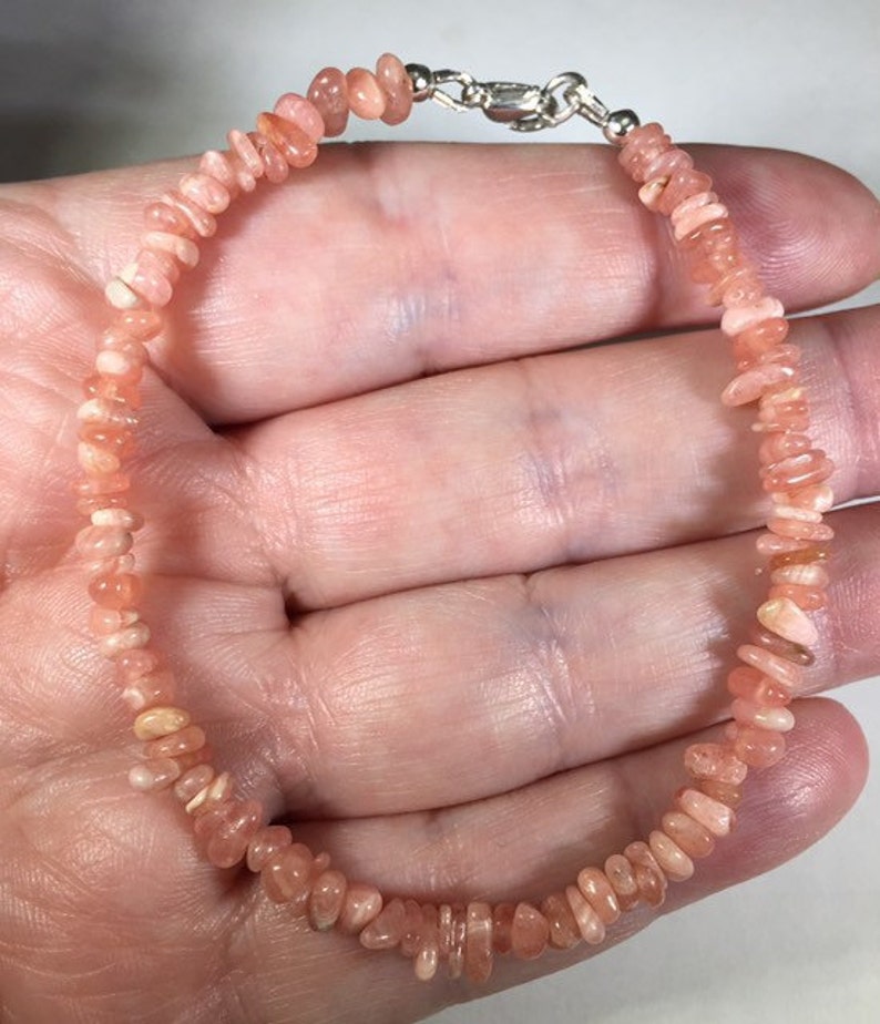 May include: A delicate bracelet featuring small, irregularly shaped, peach-colored gemstones. The stones are strung together, forming a continuous loop, and secured with a silver-colored clasp. The bracelet is designed to be worn around the wrist.