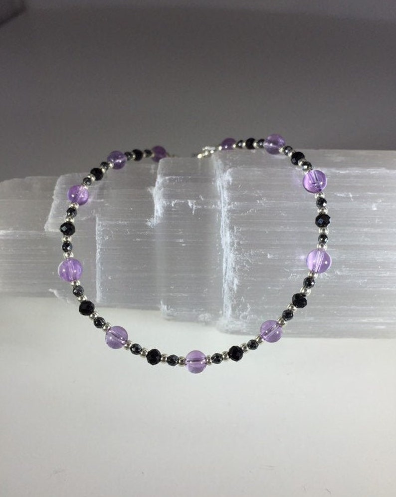 May include: A delicate bracelet made with alternating amethyst and black beads. The bracelet is strung on a silver chain.