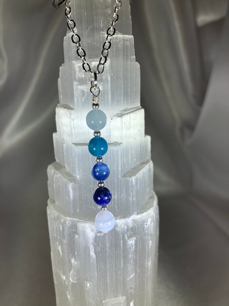 May include: A silver chain necklace with a pendant of graduated blue gemstone beads. The beads range in color from light blue to deep indigo. The necklace is displayed against a white, textured background.