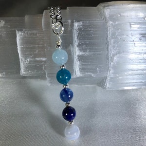 May include: A silver chain necklace with a pendant featuring a gradient of blue and white gemstone beads. The beads range in color from light blue to deep navy. The necklace is displayed against a white, textured background.