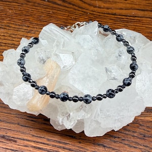 May include: A black beaded bracelet with snowflake obsidian stones and a silver clasp rests on a white crystal cluster. The bracelet is composed of small, round beads. The background is a wooden surface.