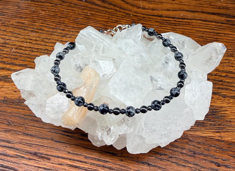 May include: A black beaded bracelet with snowflake obsidian stones and a silver clasp rests on a white crystal cluster. The bracelet is composed of small, round beads. The background is a wooden surface.