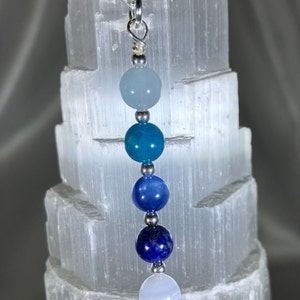 May include: A silver chain necklace featuring a pendant with a gradient of blue gemstone beads. The beads range in color from light blue to deep indigo. The necklace is displayed against a textured white background.