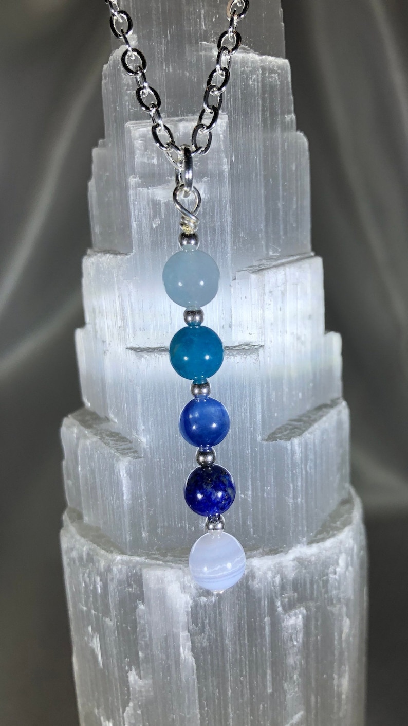 May include: A silver chain necklace featuring a pendant with a gradient of blue gemstone beads. The beads range in color from light blue to deep indigo. The necklace is displayed against a textured white background.