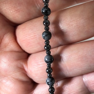May include: A beaded bracelet featuring a mix of black and gray snowflake obsidian and hematite beads. The bracelet has a silver-colored charm at the end. The beads are strung together on a dark cord.