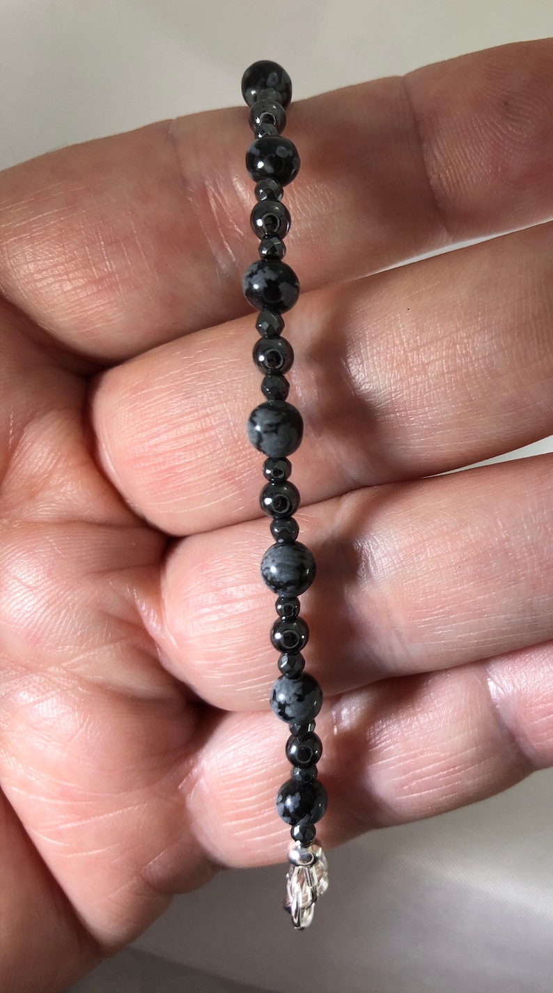 May include: A beaded bracelet featuring a mix of black and gray snowflake obsidian and hematite beads. The bracelet has a silver-colored charm at the end. The beads are strung together on a dark cord.