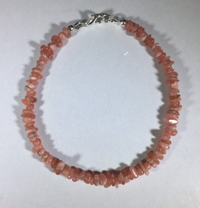 May include: A delicate bracelet featuring small, irregularly shaped, peach-colored gemstones. The stones are strung together, creating a continuous loop. The bracelet is secured with a silver-colored clasp.