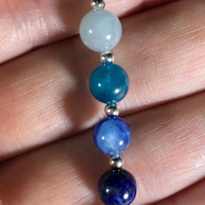 May include: A silver necklace featuring a series of colorful gemstone beads. The beads are in shades of blue, including aquamarine, teal, and lapis lazuli. The necklace is suspended from a silver chain.
