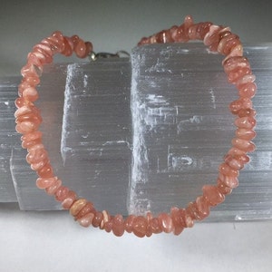 May include: A bracelet made of small, irregular, peach-colored stones. The stones are strung together with a silver-colored clasp. The bracelet is displayed against a white, textured background.
