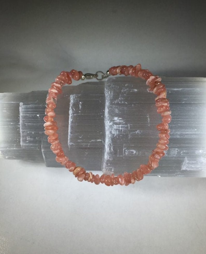 May include: A delicate bracelet composed of small, irregularly shaped, light orange-pink gemstones. The bracelet is strung together with a silver-colored clasp. The gemstones are arranged in a circular pattern. The background is a light gray, textured surface.