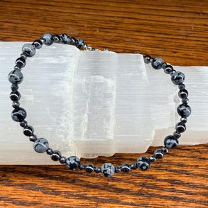 May include: A beaded necklace featuring black and gray snowflake obsidian and hematite beads. The necklace is strung on a silver-colored chain and rests on a white crystal formation, set against a wooden surface.