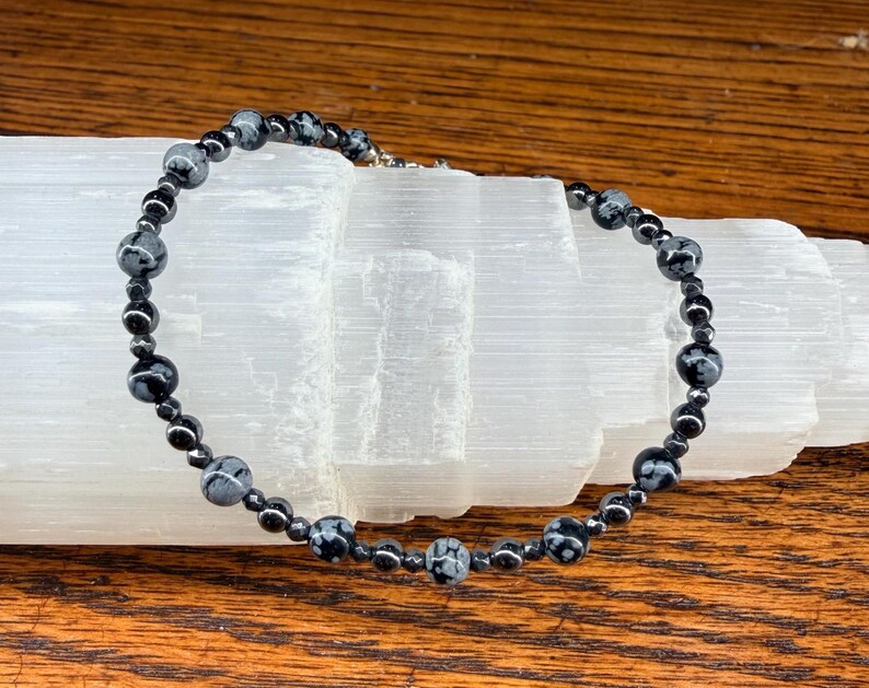 May include: A beaded necklace featuring black and gray snowflake obsidian and hematite beads. The necklace is strung on a silver-colored chain and rests on a white crystal formation, set against a wooden surface.
