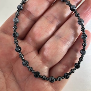 May include: A beaded bracelet featuring a mix of black and gray snowflake obsidian and hematite beads. The bracelet is strung on a flexible cord, with the beads varying in size and shape. The bracelet is shown on a person's hand.