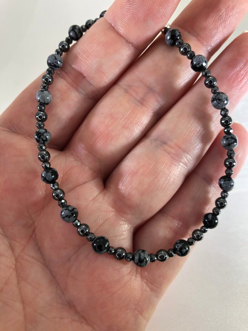 May include: A beaded bracelet featuring a mix of black and gray snowflake obsidian and hematite beads. The bracelet is strung on a flexible cord, with the beads varying in size and shape. The bracelet is shown on a person's hand.