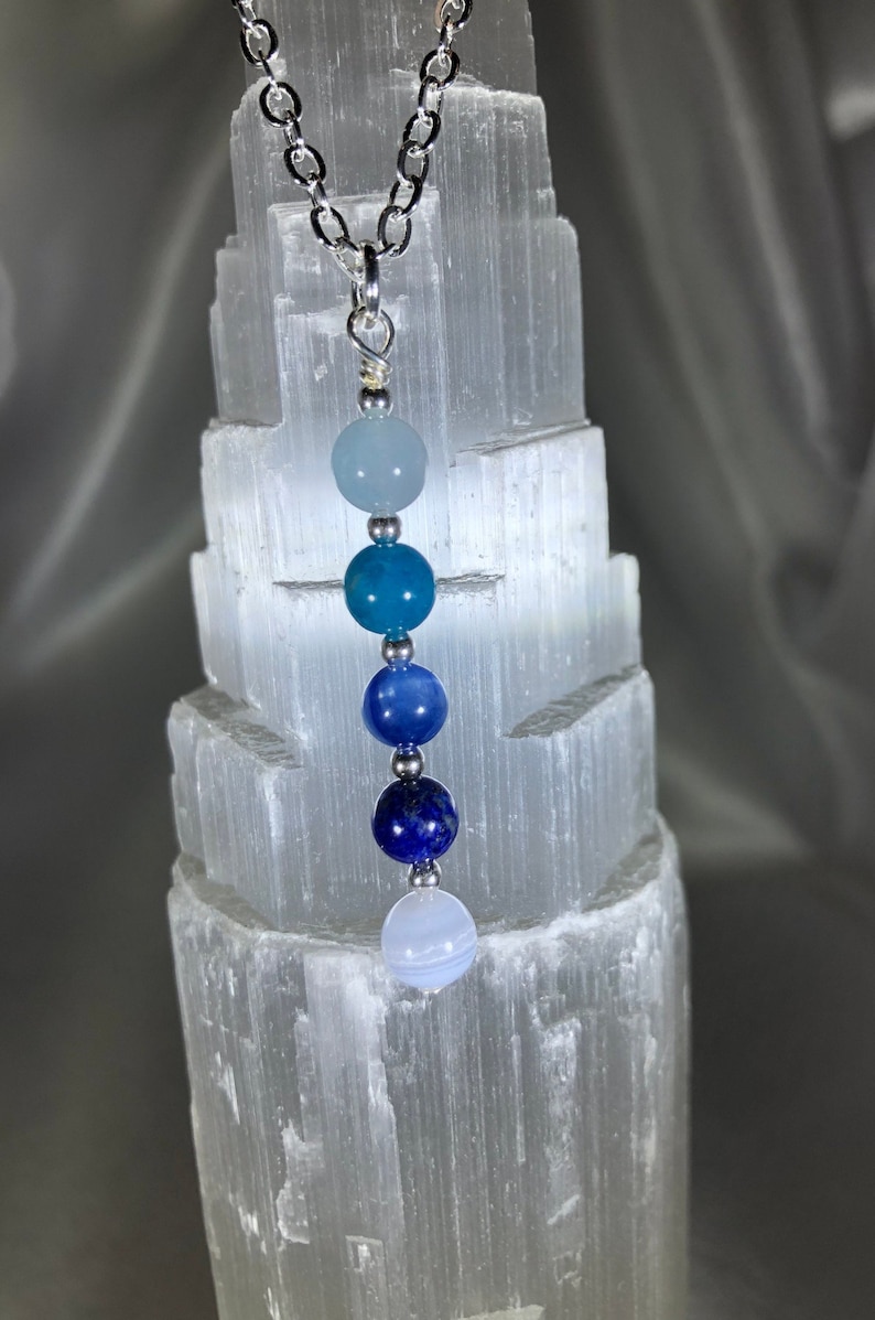 May include: A silver chain necklace featuring a series of blue gemstone beads in varying shades. The beads are round and strung together, creating a gradient effect from light to dark blue. The necklace is displayed against a textured white background.