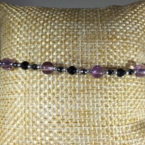 May include: A bracelet made with black onyx and amethyst beads. The bracelet is on a beige fabric background.