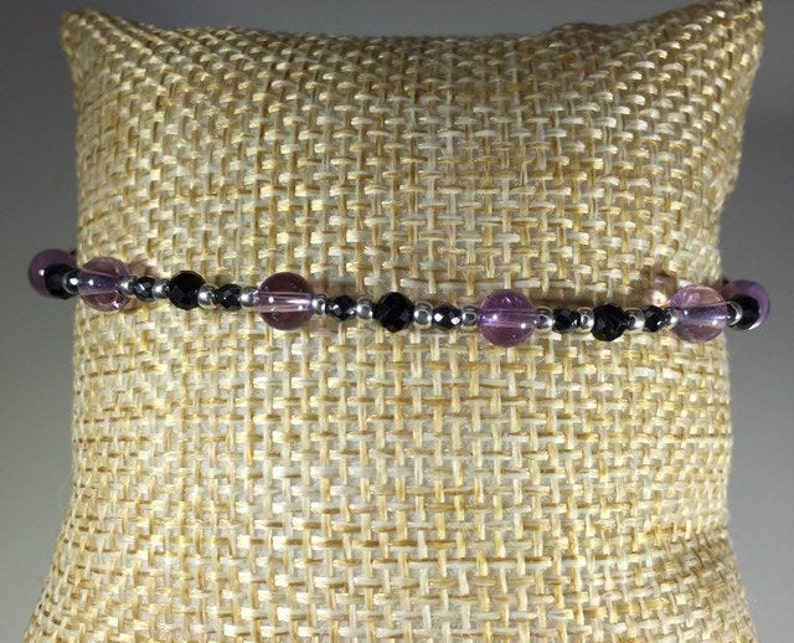 May include: A bracelet made with black onyx and amethyst beads. The bracelet is on a beige fabric background.