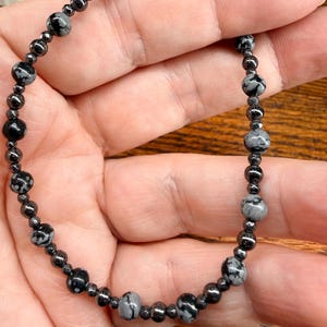 May include: A beaded bracelet featuring a mix of black and gray snowflake obsidian and small, dark hematite beads. The bracelet is strung on a dark cord and is held in a hand. The bracelet is a jewelry item.