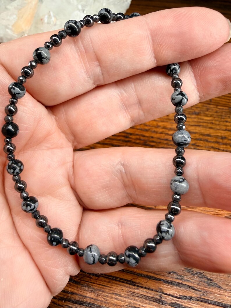 May include: A beaded bracelet featuring a mix of black and gray snowflake obsidian and small, dark hematite beads. The bracelet is strung on a dark cord and is held in a hand. The bracelet is a jewelry item.