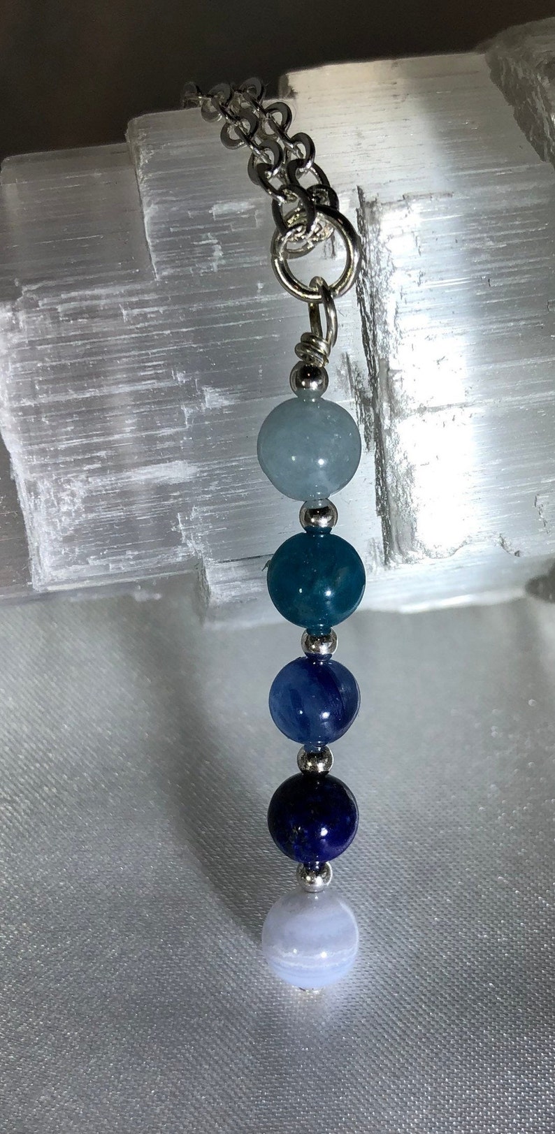 May include: A silver chain necklace featuring a series of colorful gemstone beads. The beads are in shades of blue, ranging from light blue to deep indigo, with silver accents. The necklace is displayed against a textured white background.