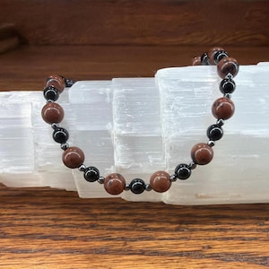 May include: A beaded bracelet featuring alternating round brown and black beads, accented with smaller metallic grey spacer beads. The beads are arranged in a repeating pattern along the circular strand, creating a consistent design throughout the piece.