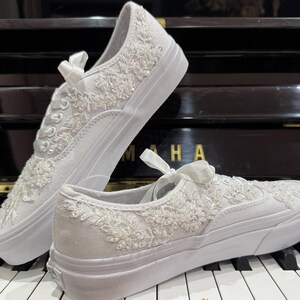 May include: Two white canvas sneakers with intricate floral embroidery and embellishments. The shoes feature a classic low-top design with white laces and a white rubber sole. A satin bow adorns the laces. The shoes are displayed on a piano keyboard.