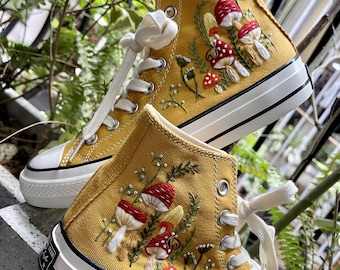 Può includere: Un paio di scarpe da ginnastica alte in tela gialla con funghi e fiori ricamati. Le scarpe hanno lacci bianchi e una suola in gomma bianca.