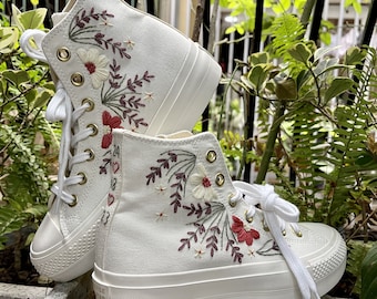 May include: White high-top sneakers with floral embroidery in red, cream, and burgundy. The canvas shoes feature white laces, a thick white sole, and gold eyelets. The design includes embroidered flowers and leaves.