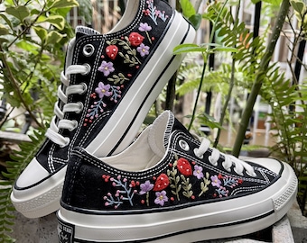 Può includere: Sneakers in tela nera con suole bianche e fiori e fragole ricamate in rosso, verde e viola.