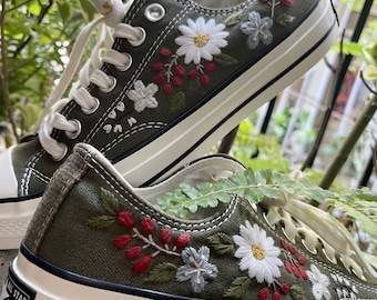 May include: Olive green canvas sneakers with white soles and laces. The sneakers are adorned with embroidered floral designs in white, red, and gray. The design includes daisies and other flowers.