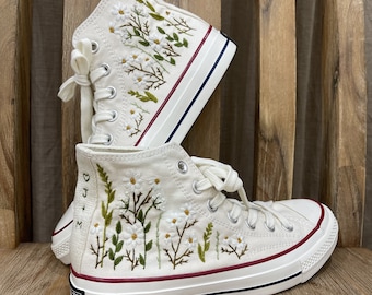 May include: White high-top canvas sneakers with embroidered white flowers and green leaves. The sneakers have a red stripe around the top and a black stripe around the sole. The text "Love + X" is embroidered on the side of the shoe.