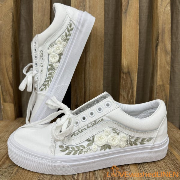 Può includere: Un paio di scarpe da ginnastica bianche in tela con fiori e vegetazione ricamate. Le scarpe hanno il testo "Karon & Selina" e "04.01.2025" ricamato.