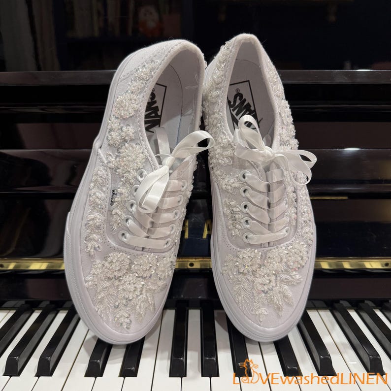 May include: A pair of white Vans sneakers, heavily embellished with floral embroidery and beadwork. The shoes feature white satin ribbon laces and are set against a piano keyboard. The Vans logo is visible on the side.