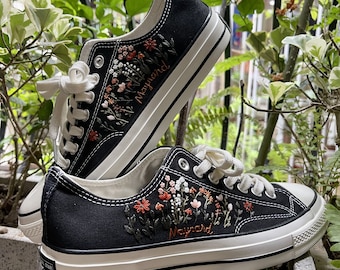 May include: Black canvas sneakers with white laces and soles. The sneakers feature embroidered floral designs in shades of pink, orange, and white. The name "Maynard" is embroidered on the side of each shoe.