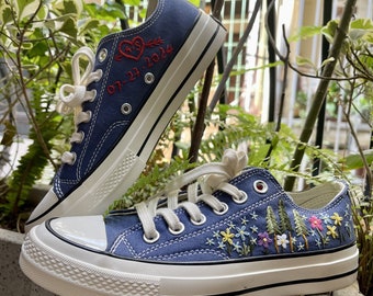 Puede incluir: Un par de zapatillas de lona azules con suelas blancas. Un zapato tiene bordado en rojo un corazón con una flecha y la fecha 07-27-2024. El otro zapato tiene bordado de flores y árboles coloridos.