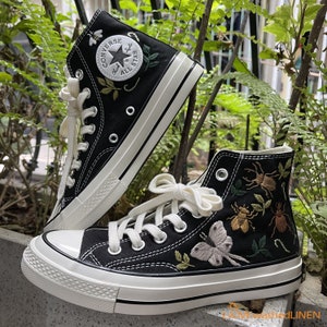 May include: A pair of black high-top canvas sneakers with white stitching and a white rubber sole. The sneakers are decorated with embroidered insects and flowers. The left sneaker has the Converse logo and the text "Chuck Taylor All Star".