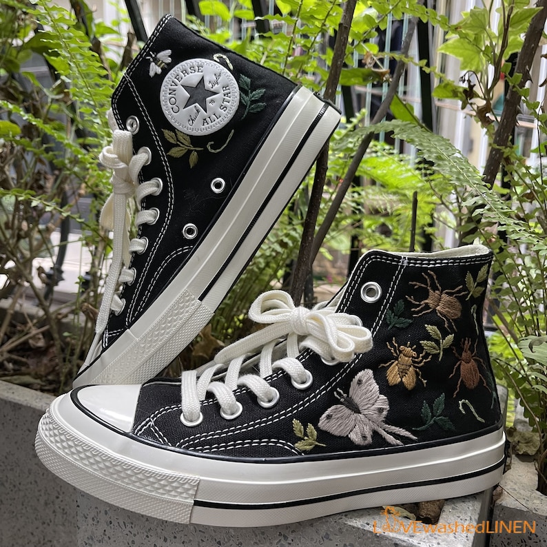 May include: A pair of black high-top canvas sneakers with white stitching and a white rubber sole. The sneakers are decorated with embroidered insects and flowers. The left sneaker has the Converse logo and the text "Chuck Taylor All Star".