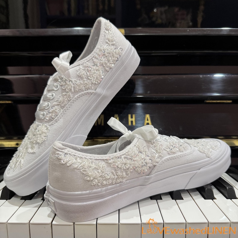 May include: Two white canvas sneakers with intricate floral embroidery and embellishments. The shoes feature a classic low-top design with white laces and a white rubber sole. A satin bow adorns the laces. The shoes are displayed on a piano keyboard.